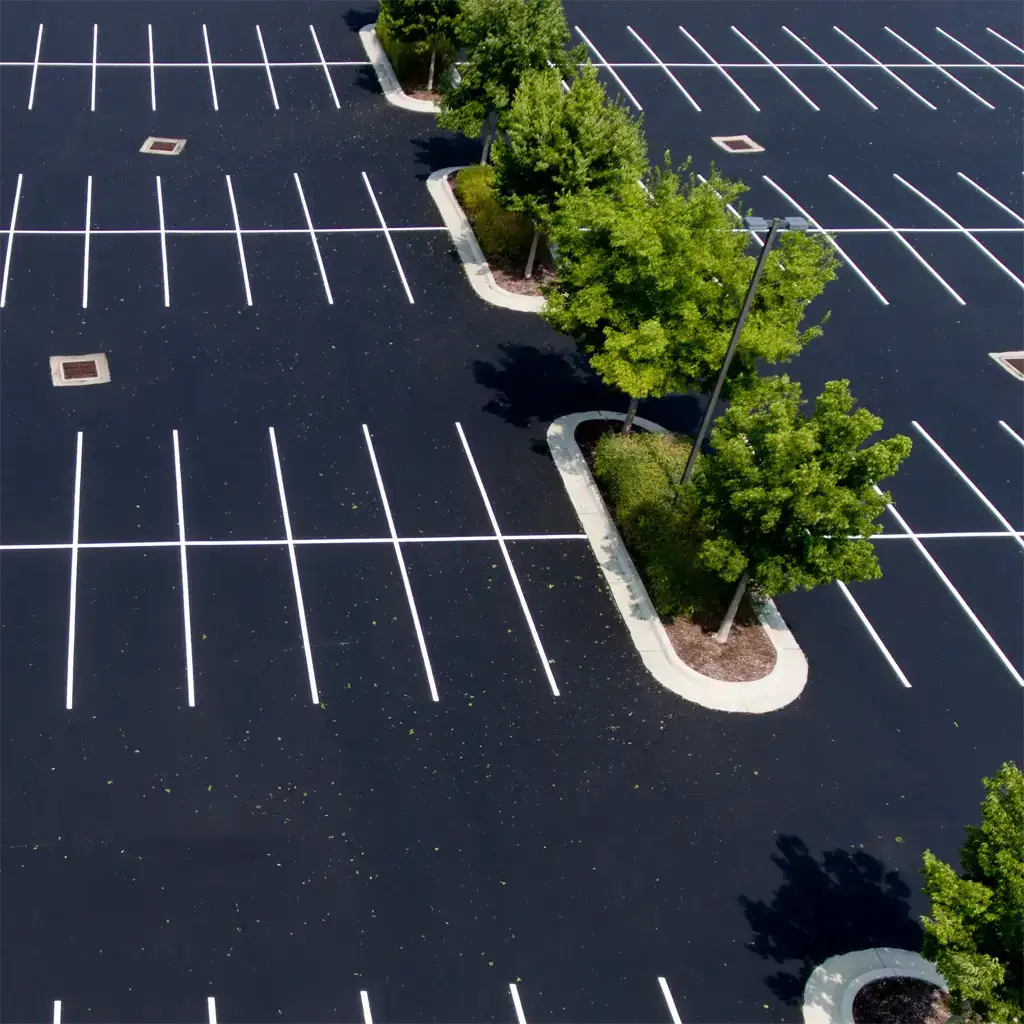nj-asphalt-paved-parking-lot Aerial view of an empty parking lot with rows of marked spaces separated by landscaped islands with trees.